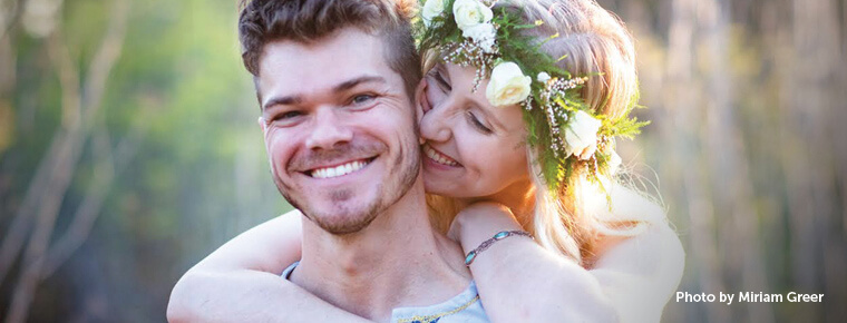 A man smiles at the camera while a woman wearing a flower wreath on her head hugs him from behind