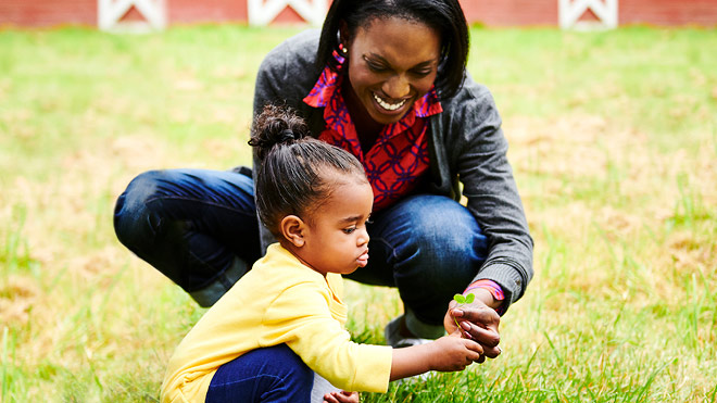 A toddler squats in the grass, looking at a clover held by her smiling mother.