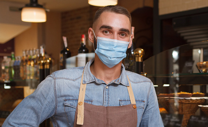 Cafe worker wearing surgical mask over his nose and mouth standing in front of the counter