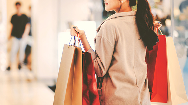 A woman wearing a tan coat, carrying shopping bags walks through a mall.