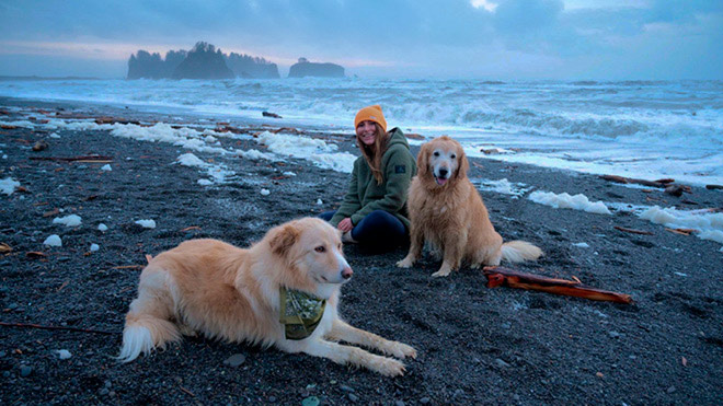 A woman dressed in warm clothes sits on a Pacific Northwest beach with two dogs