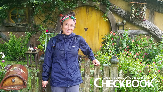 Smiling woman in a blue windbreaker stands in a garden in front of cabin with a low, round wooden door. Logo in the lower right reads Puget Sound Consumers' Checkbook.