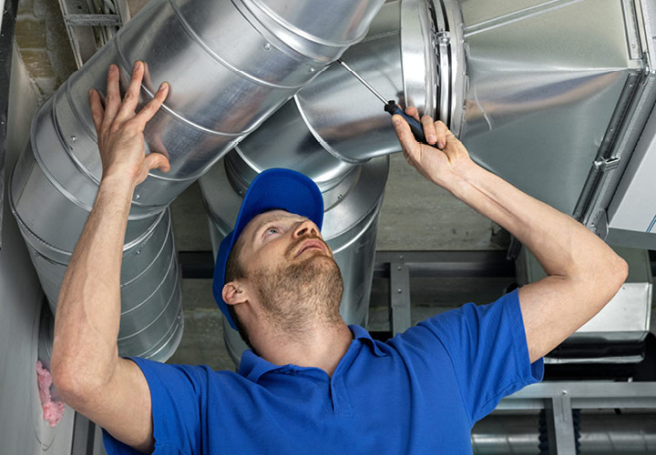 Individual in royal blue collared t-shirt and royal blue ballcap wields a screwdriver while inspecting ductwork in their home.