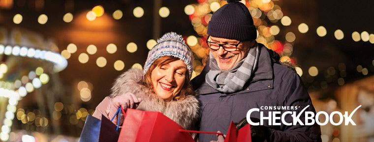 Couple standing shoulder to shoulder together outside, dressed in warm winter clothes, smiling and looking down inside of a red shopping bag, with holiday lights visible in the background. "Consumers' Checkbook" logo in the bottom right corner.