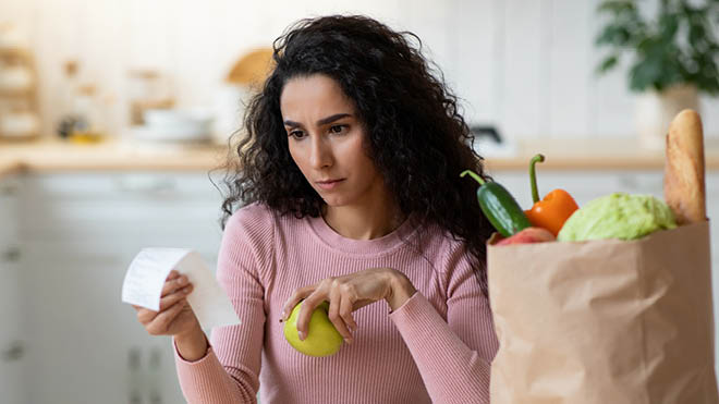 A woman sits at her kitchen table reading a receipt she is holding in one hand. She is holding an apple in the other hand. A paper bag of groceries is on the table next to her.