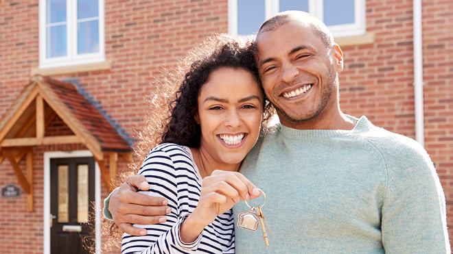 Image of a young, smiling couple standing in front of a brick home with one of the individuals holding house keys in their hand.