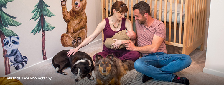 Mother and father sitting on the floor in front of a baby crib holding and looking at their newborn baby, with a black- and white-colored dog as well as a brown- and grey-colored dog laying down on the floor next to them. Friendly depictions of a raccoon, bear, and forest are painted on the walls of the room. "Miranda Jade Photography" photo credit is displayed in text in bottom left hand corner of image.