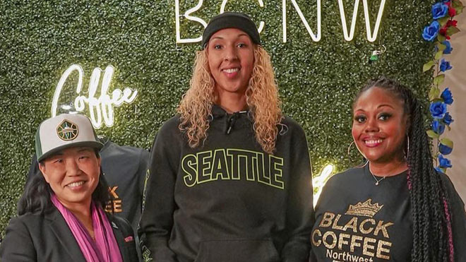 Three women standing together in front of a green, leaf-covered wall with a neon sign that says "Coffee" on it. The woman on the left is Intentionalist CEO and founder Laura Clise, wearing a Seattle Sonics hat. The woman in the middle is Seattle Storm Center Mercedes Russell, wearing a sweatshirt that says "Seattle." The woman on the right is Black Coffee Northwest co-owner DarNesha Weary, wearing a "Black Coffee Northwest" t-shirt.