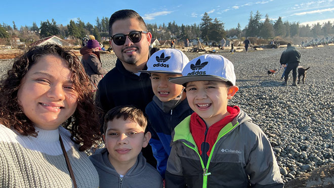Image of BECU employee Marybeth with her husband, Jose, and their three children, (from left) Ayden, Derek and Nathan smiling for a photo outside. A rocky beach is pictured in the background.
