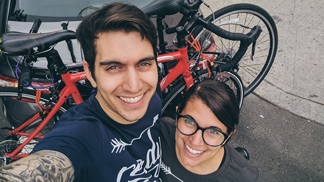 A couple smiling, taking a selfie. A pair of bicycles are on a car-mounted bike rack in the background.