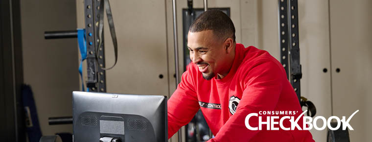 A man is smiling and riding a stationary bike indoors. In the background is fitness equipment. He is looking at a screen while riding the bike. Both of his hands are on the handle bars and he is wearing a red crewneck sweater. The photo has a Consumers' Checkbook logo and the bottom right hand corner.