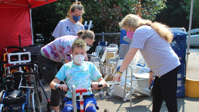 A young boy is sitting on an adaptive bike, while three individuals help to make sure the bike fits correctly. All three women are looking at the bike. Two are wearing gray shirts and one is wearing a tie dye shirt. In the background is other assistive devices such as wheelchairs, walkers and other equipment. They are outside under a red canopy tent. Behind the group are trees and a couple parked cars.