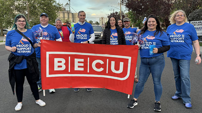 A group of BECU members participating in the Spokane Lilac Parade. They are holding a red BECU banner, wearing blue Veterans t-shirts and smiling proudly.