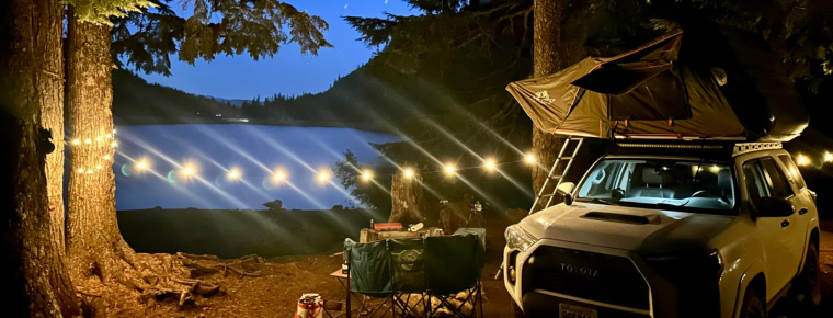 A campsite by a lake at twilight with a vehicle and rooftop tent. Chairs and a table are set up near the vehicle, and string lights are hung between trees.