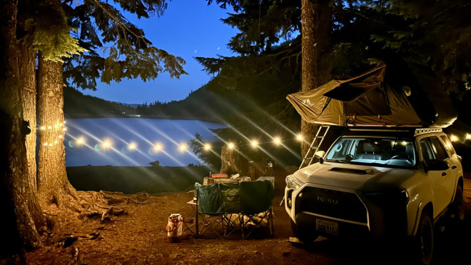 A campsite by a lake at twilight with a vehicle and rooftop tent. Chairs and a table are set up near the vehicle, and string lights are hung between trees.