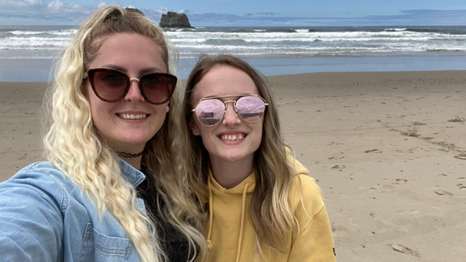 A mother and daughter standing on a sandy beach, both smiling and wearing sunglasses. The ocean, white waves, blue sky and a large rock are visible in the background. 