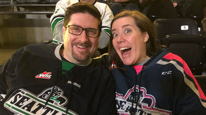 A couple at a hockey game, wearing Seattle Thunderbirds jerseys. The man, on the left, is smiling and wearing glasses. The woman, on the right, looks excited, posing with her mouth open. The two are seated in a hockey arena setting. 