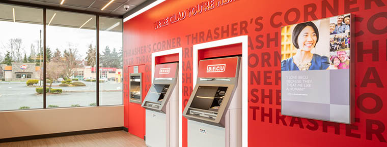 Two ATMs located inside a BECU neighborhood financial center. The ATMs are placed on a red BECU branded wall. There is a BECU branded photograph on the wall as well. On the left is a window showing an outside parking setting. 