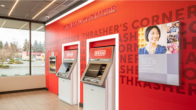 Two ATMs located inside a BECU neighborhood financial center. The ATMs are placed on a red BECU branded wall. There is a BECU branded photograph on the wall as well. On the left is a window showing an outside parking setting. 