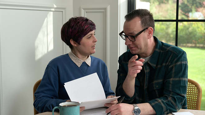 A man and woman are sitting close together at a table having a conversation. They appear relaxed and their expressions are calm. A cup of coffee and some papers are on the table. They are sitting in front of a white wall with picture molding and a window with a green lawn outside. Sun is shining in the window. The woman is looking at the man and talking while holding a stack of papers. The top few sheets are flipped up so they can look at a page in the stack. The man is looking down at the papers, gesturing with one hand and writing on a piece of paper with the other. 