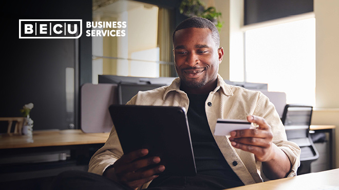 A man looking at a tablet while holding his business credit card. BECU Business Services logo.