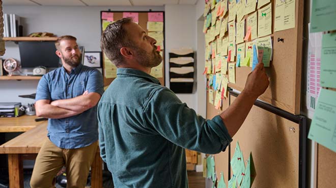 Image shows two guys looking at a board covered in Post-Its.