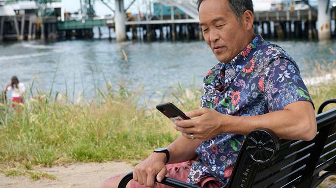 Image is of a man sitting outside on a park bench and looking at his phone. There is a body of water and a bridge in the background.