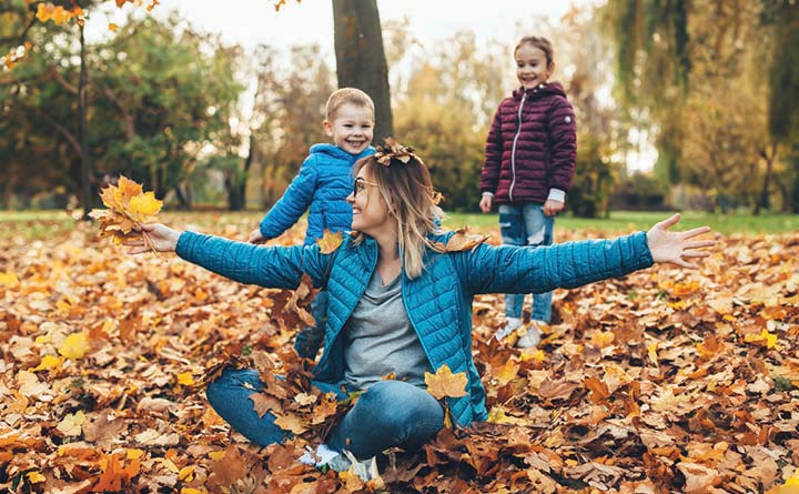 Image shows a smiling woman sitting cross-legged in a pile of dry leaves. The season is autumn. The woman has her arms outstretched and she is looking to the side and behind her, where two young children in jackets are standing and smiling.