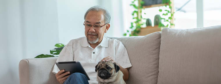 Older man sitting on a couch, looking at tablet and petting the dog seated next to him.