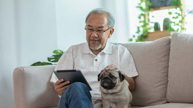 Older man sitting on a couch, looking at tablet and petting the dog seated next to him.