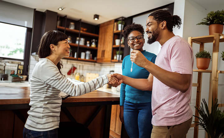 Image is of three young people who appear to be in their mid to late 20s standing facing each other in a kitchen. The person in the left side of the image is smiling and offering a handshake to the person in the right side of the image. A third smiling person is in the background.