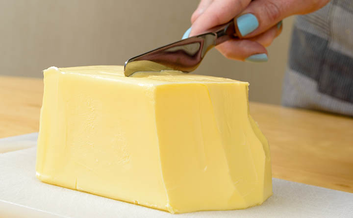 A hand with blue nail polish cuts into a stick of butter with a butter knife.
