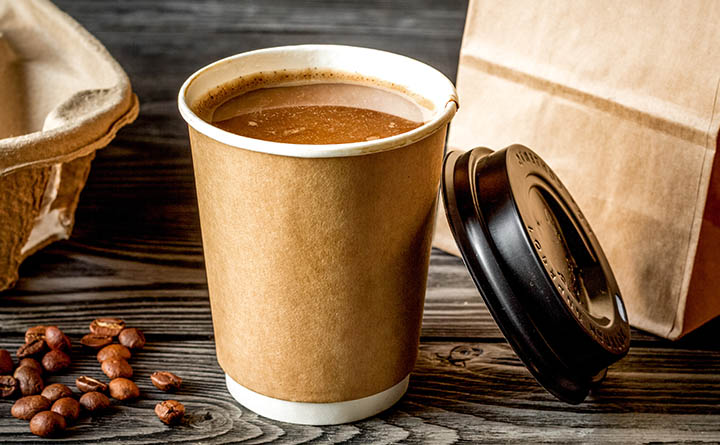 A full cup of coffee in a paper cup sits on a wood surface. A black plastic lid leans against the cup. A few coffee beans and a brown paper bag of coffee sit on either side of the cup. The corner of a carboard carrying tray is visible.