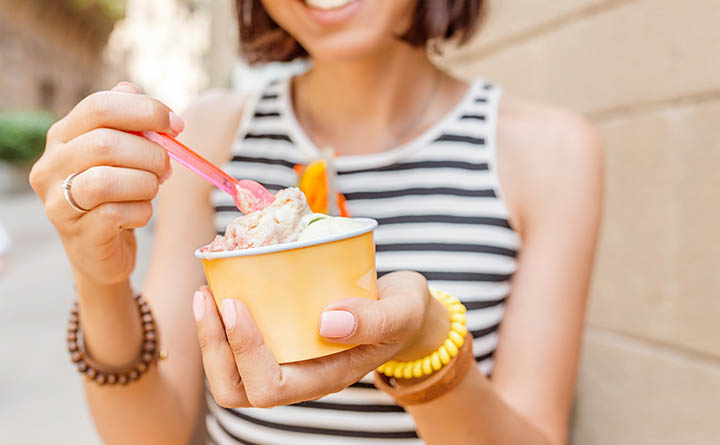 A woman in a striped tank top dips a pink plastic spoon into a yellow cardboard dish of ice cream with sprinkles.