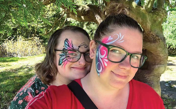 BECU employee Amanda G and her partner pose for a selfie outdoors. The two have butterfly and floral face paintings. Both are wearing glasses and there is a tree in the background behind him. 