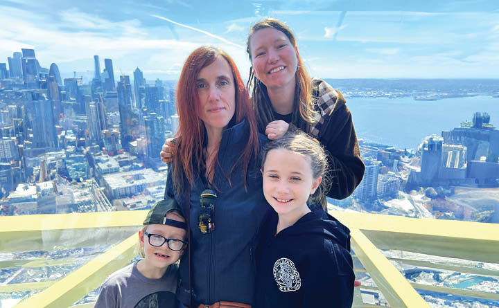 BECU employee Liz H poses with her family (two young kids and her partner) for a photo with a city landscape behind them.