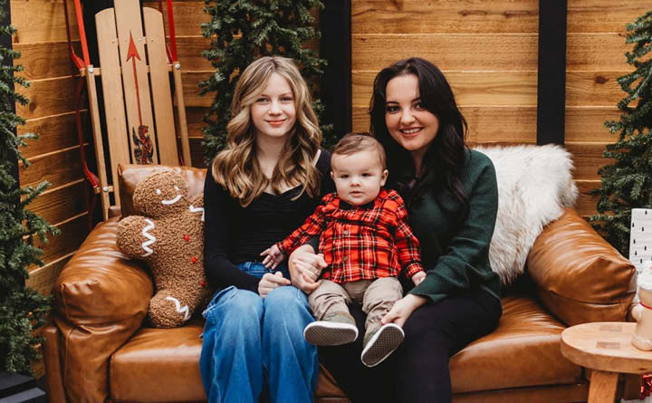 BECU employee Nakia C poses for a family photo with her son and daughter during the holiday season. The son is seated in between her and her daughter on their laps. The family is seated on a couch. There is a gingerbread man pillow on the couch, a sled in the background and Christmas trees. 