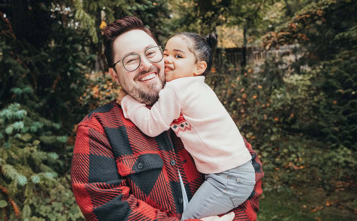 BECU employee Tanner A smiles and holds his daughter. Her arms are wrapped around him. He is wearing a red and black checkered flannel, and she is wearing a pink sweater. There are trees and flowers in the background.
