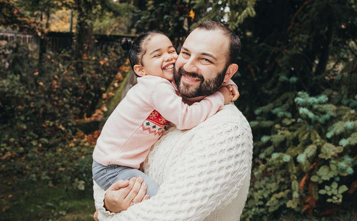 BECU employee Tanner A's partner smiles and holds his daughter for an outdoor photo. Her arms are wrapped around him and she hugs him. The daughter is wearing a pink shirt and he is wearing a cream sweater. There is trees and flowers in the background. 