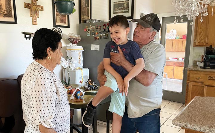 In a kitchen, a grandfather holds his young grandson, as the grandmother stands nearby. The grandson is smiling and laughing. In the background are framed photos, a refrigerator and wooden kitchen cabinets. 