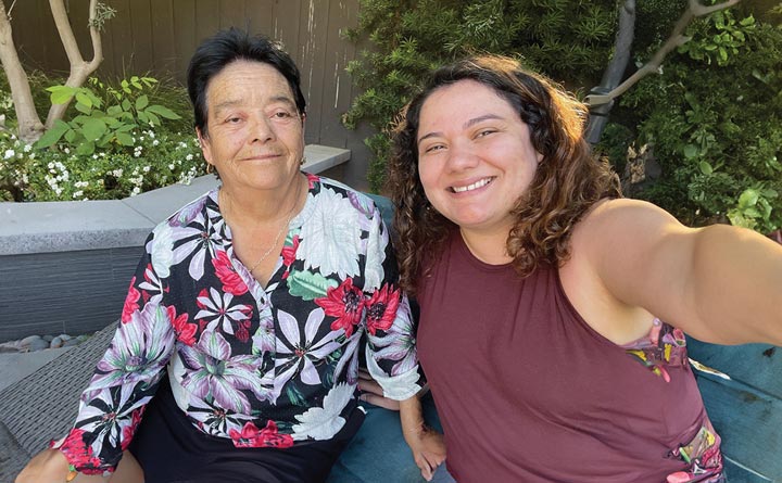 BECU employee, Marybeth R. (right) and her grandmother (left) pose for a selfie together on a bench outside. There are some plants in the background and a wooden fence.