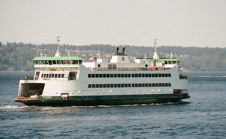 A passenger ferry sailing nearing Bainbridge Island in Washington.  The ferry is white with green accent colors. In the background are trees, homes and the shoreline, capturing gray skies. 