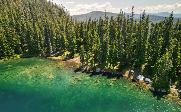 An aerial view of a Lake Kachess in Washington, nestled among tall trees. The water is a bright green color. In the background are hills, clouds and blueish gray skies. 