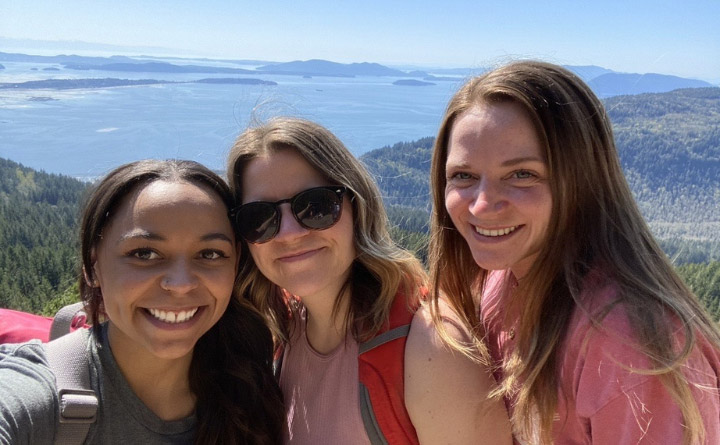 Three women smiling and posing for a selfie at the peak of Oyster Dome trail, capturing a mountain and bay-water view.