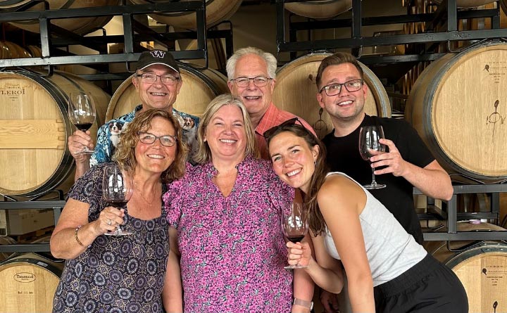 A group of six people smiling and holding wine glasses while posing for a photo. There are wine barrels in the background. 