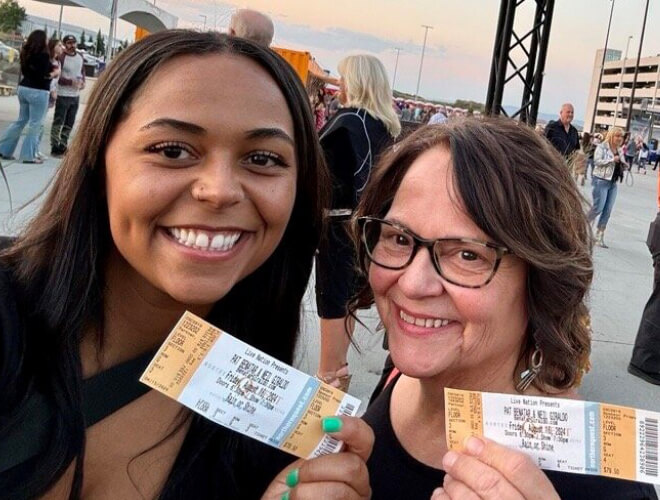 Two women hold their tickets at a lively outdoor concert event. In the background are people and a sunset view. 