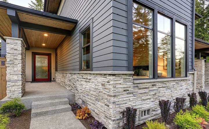 A close up of a home with stone veneer on the lower third. The home is gray and has a large window in the front. There is also stone veneer on a column close to the front entryway. Steps lead up to a front door with patio lights on. There is landscaping along the base of the home.