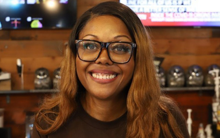 A woman in glasses is smiling at the camera. Behind her is a wood wall with two tv screens.