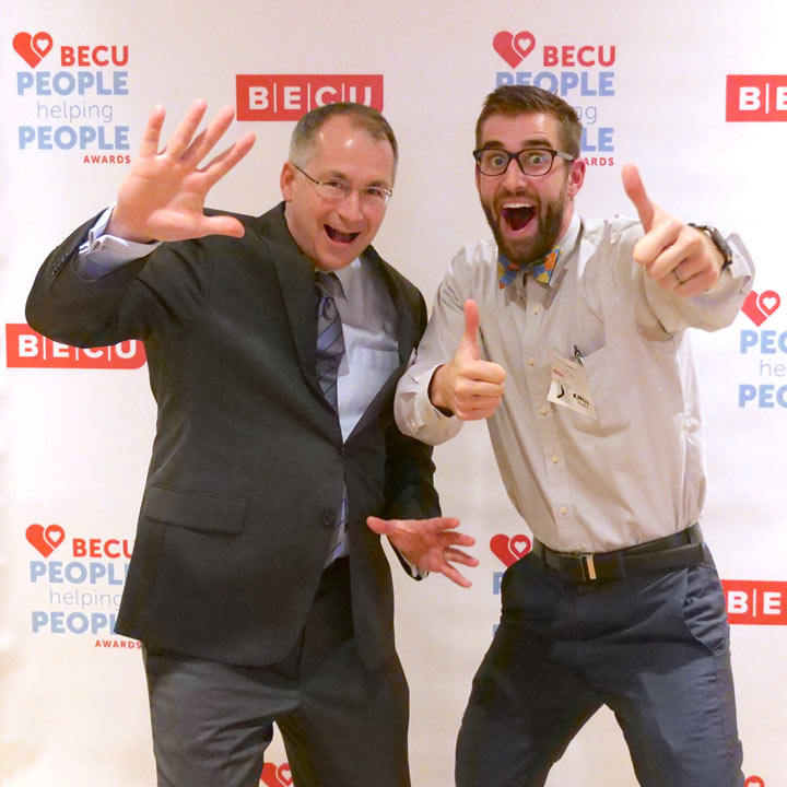 Benson Porter smiles and waves excitedly with a smiling award recipient giving two thumbs up to the camera. They are standing in front of a backdrop that says BECU and People Helping People.
