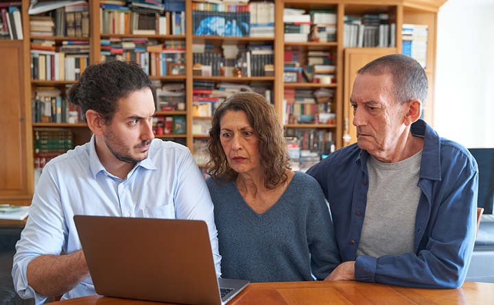 Image shows a seated middle-aged female and man looking at laptop with shocked expressions. Next to them is a younger man who is pointing out something on the computer screen.